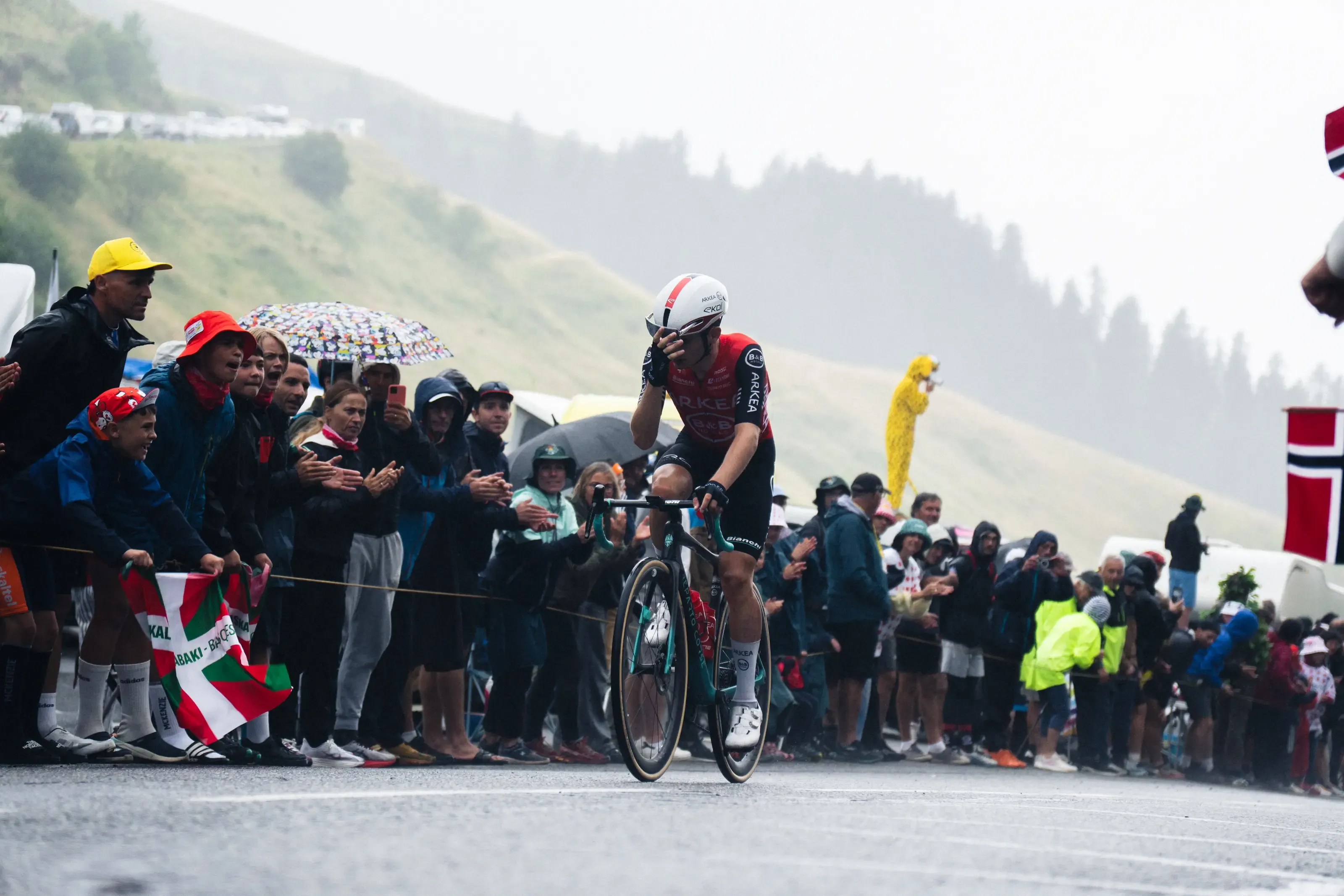 Une photographie de course cycliste professionnelle en haute montagne, se déroulant sous une pluie battante et un ciel gris brumeux. Au centre, un cycliste solitaire de l'équipe Arkea, portant un maillot rouge et noir et un casque blanc, roule sur une route mouillée, s'essuyant le front ou se tenant la tête dans un geste d'épuisement ou de déception. Sa main gauche est sur son front. La foule est alignée sur le côté gauche de la route derrière une corde de sécurité, acclamant le cycliste malgré les conditions météorologiques. Les spectateurs sont emmitouflés dans des imperméables et tiennent des parapluies. Un enfant tient un drapeau basque. À l'arrière-plan, la route serpente sur une colline brumeuse et boisée. À droite, on aperçoit un drapeau norvégien et une personne costumée en Pikachu jaune parmi la foule. L'asphalte est sombre et brillant de pluie.